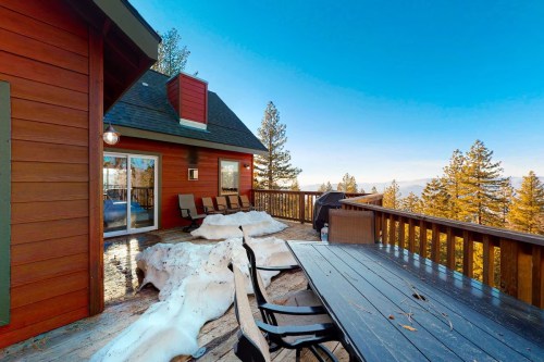 A wooden deck with chairs and a table, surrounded by snow and trees, under a clear blue sky.