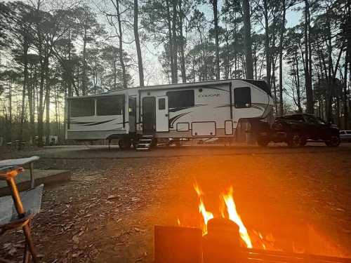 A camper parked in a wooded area at dusk, with a campfire in the foreground and trees surrounding the scene.