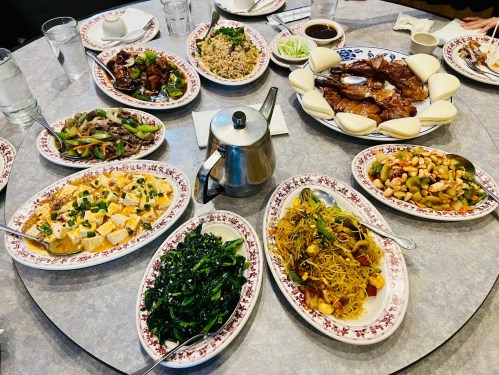 A round table set with various dishes, including meats, noodles, vegetables, and rice, alongside tea and sauces.