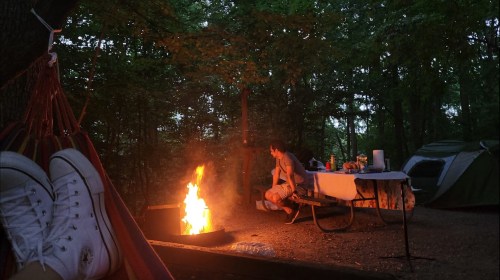 A cozy campsite at dusk, featuring a campfire, a person sitting nearby, and a tent in the background.