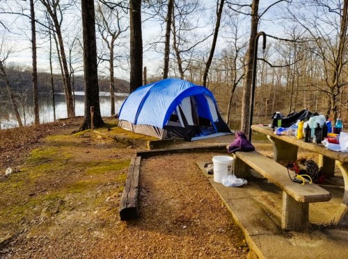 A blue tent set up near a lake, surrounded by trees and a picnic table with camping supplies scattered around.
