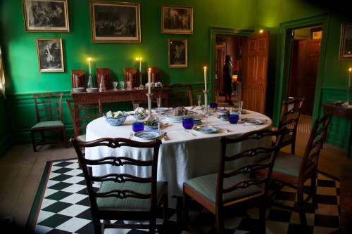 A dining room with a green wall, a round table set for dinner, and vintage decor, including candles and artwork.