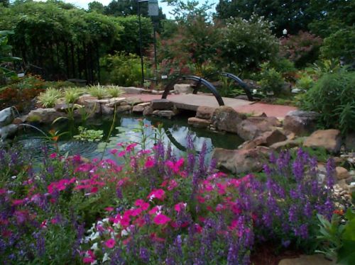 A vibrant garden with colorful flowers, a pond, and a small bridge surrounded by lush greenery.