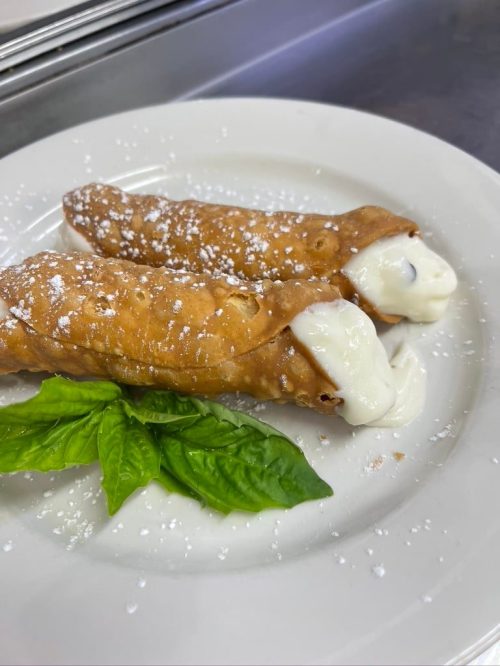 Two cannoli filled with creamy filling, dusted with powdered sugar, served on a white plate with a basil leaf.
