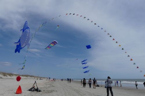 Colorful kites flying in the sky over a beach, with people walking along the shore under a cloudy sky.