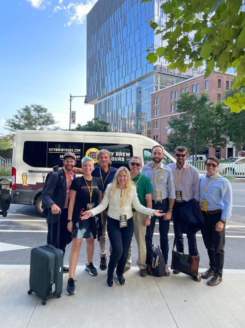 A group of eight people poses cheerfully in front of a van, with a modern building in the background.