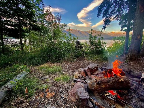 A cozy campfire surrounded by trees, with a scenic view of mountains and a lake under a colorful sky.