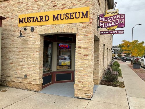 Exterior of the National Mustard Museum, featuring a sign and an open door, with a brick facade and nearby trees.