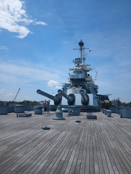 A battleship's deck with large cannons, under a blue sky with scattered clouds and a view of the horizon.