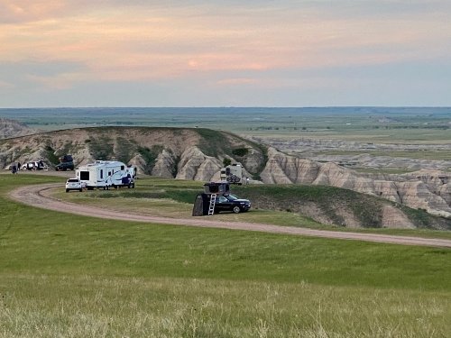 A scenic view of a winding road with RVs parked on a grassy hill overlooking rugged terrain and a colorful sky.