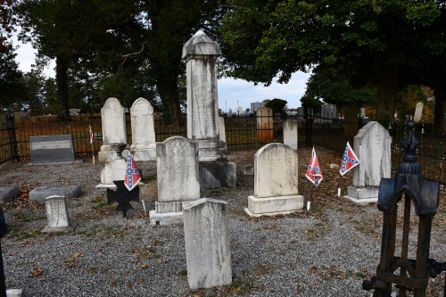 Graveyard with several headstones and Confederate flags, surrounded by a black iron fence and trees.