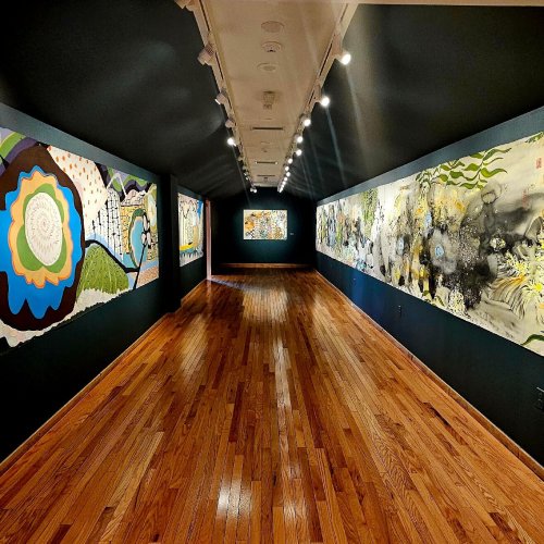 A long, narrow art gallery with wooden floors and colorful wall murals on both sides, illuminated by overhead lights.