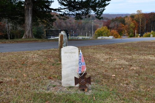 Grave marker labeled "Unknown" with a Confederate flag and cross, set in a grassy area with trees in the background.