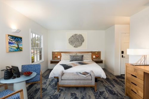 A modern hotel room featuring a queen bed, seating area, artwork, and natural light from large windows.