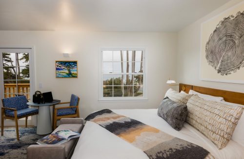 Cozy bedroom with a large bed, artwork, a small table, and a view of trees and the ocean through a window.