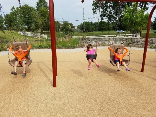 Three children in orange shirts swing on playground swings, enjoying a sunny day in a park.