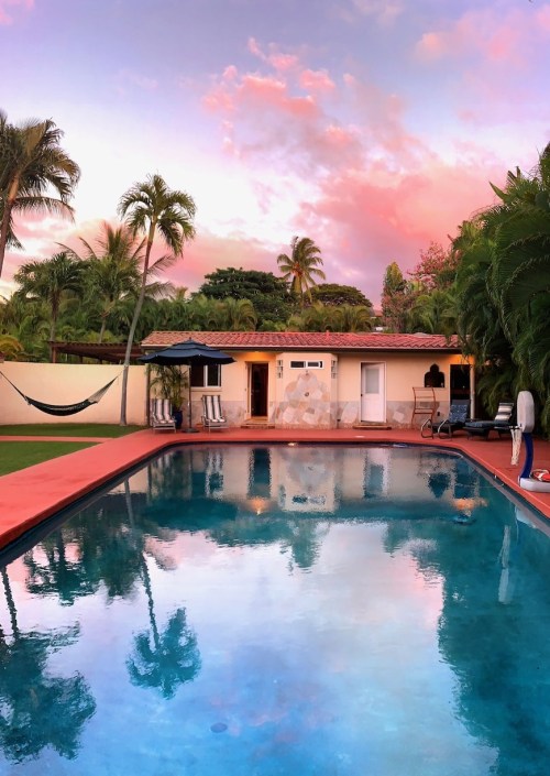 A serene pool reflects a colorful sunset, surrounded by palm trees and a cozy house.