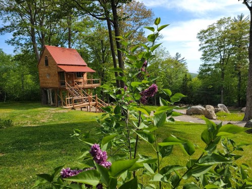 A wooden treehouse with a red roof surrounded by greenery and blooming flowers on a sunny day.