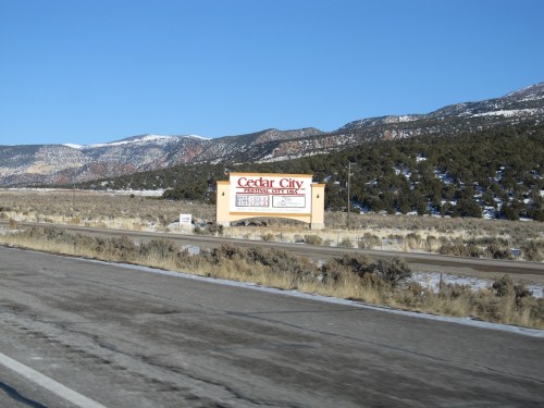 Sign for Cedar City along a highway, with mountains and clear blue sky in the background. Snow on the ground.