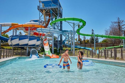 Two people walk in a pool near colorful water slides on a sunny day.