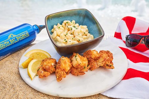 A plate with fried seafood, macaroni and cheese, lemon wedges, and a blue drink on a sandy beach. Sunglasses nearby.