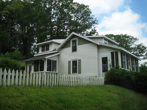 A white two-story house with a picket fence, surrounded by greenery and trees.