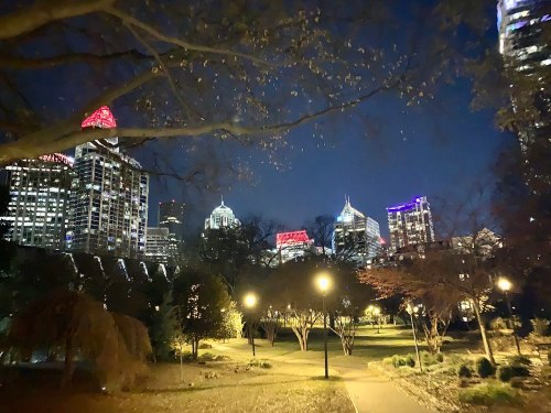 City skyline at night with illuminated buildings and trees in a park, creating a vibrant urban atmosphere.