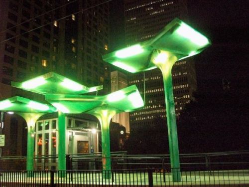 A modern bus stop illuminated with green lights, set against a city skyline at night.