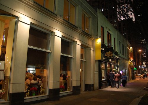 A city street at night, featuring storefronts and pedestrians walking along the sidewalk under bright lights.