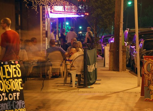 A lively street scene at night with people dining outside, colorful lights, and a sign advertising a bookstore.
