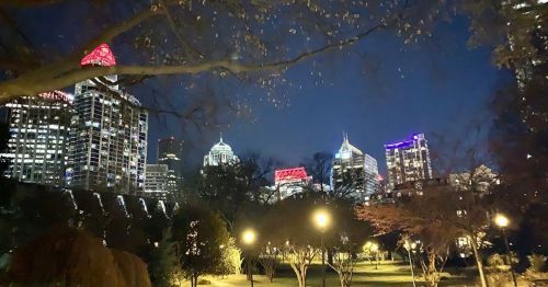 City skyline at night, featuring illuminated buildings and trees in a park, with colorful lights on skyscrapers.
