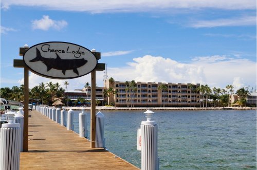 Sign for Cheeca Lodge on a wooden pier with a view of a hotel and palm trees under a blue sky.
