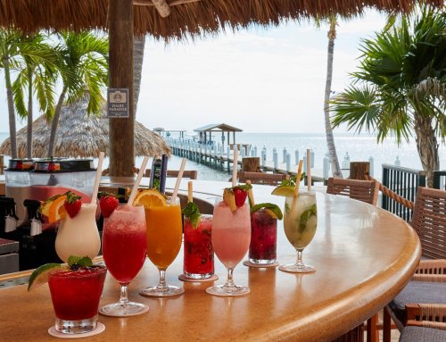 A tropical bar with colorful cocktails lined up on the counter, overlooking a serene waterfront and palm trees.