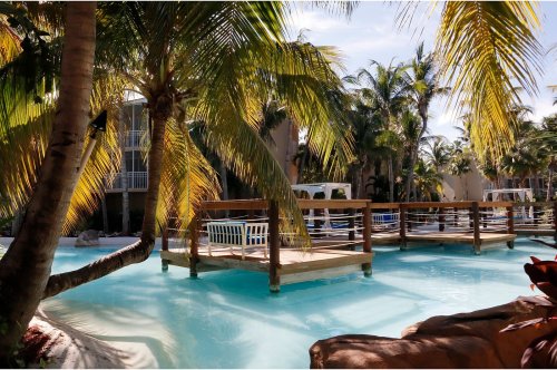 Tropical resort scene with palm trees, a wooden bridge over turquoise water, and lounge chairs in the background.