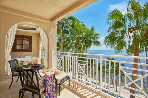 A sunny balcony with a table and chairs, overlooking palm trees and a serene ocean view.