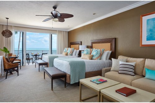 Bright hotel room with two beds, a sofa, and a balcony overlooking the ocean, featuring tropical decor and natural light.