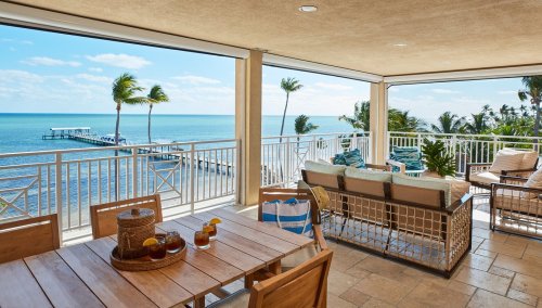 A spacious balcony with a wooden table, drinks, and a view of the ocean and palm trees under a clear blue sky.
