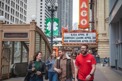 A group of people walks past a theater marquee advertising upcoming shows in a busy urban setting.