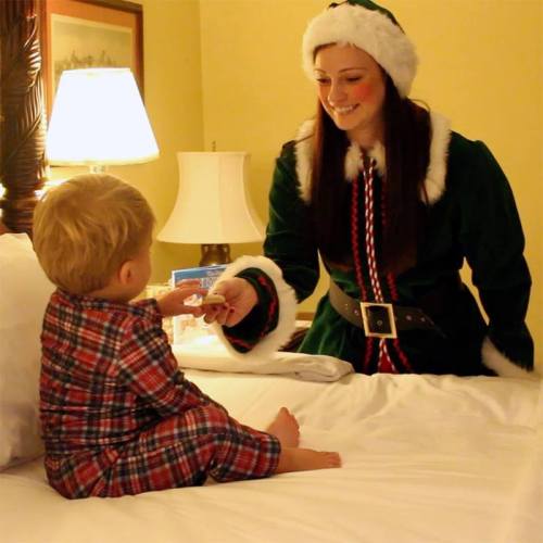 A child in pajamas sits on a bed, watching a woman in a festive outfit with a smile, surrounded by holiday decorations.