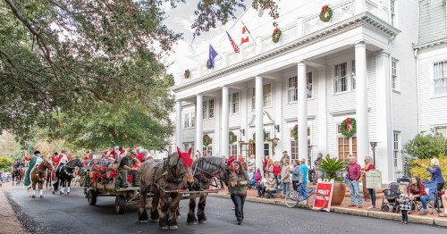 A festive parade with horses in Santa hats, passing a decorated building with onlookers and holiday decorations.