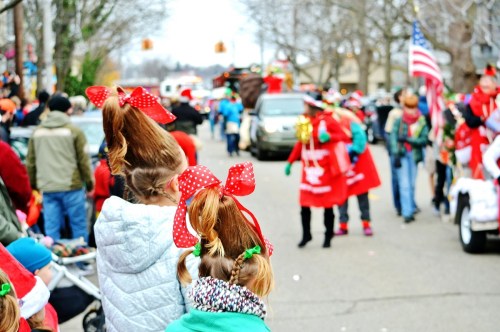 Children with festive hairstyles and bows watch a holiday parade filled with colorful costumes and decorations.
