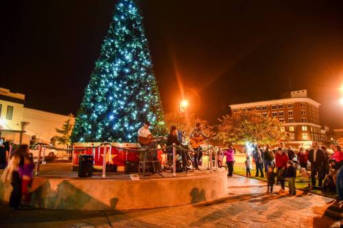 A festive scene with a large Christmas tree and musicians performing, surrounded by a crowd at night.