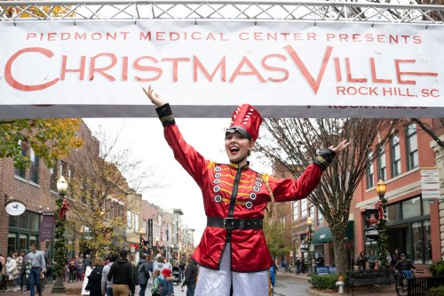 A performer in a red and gold costume poses joyfully under a "ChristmasVille" banner in a festive street scene.