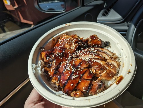 A hand holding a bowl of grilled chicken over rice, drizzled with sauce and topped with sesame seeds.