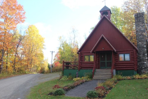 A red log cabin with a stone chimney, surrounded by colorful autumn trees and a gravel road.
