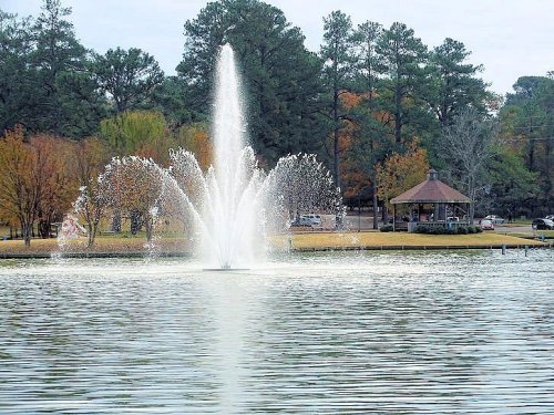 A fountain sprays water in a serene lake, surrounded by trees and a gazebo in the background.