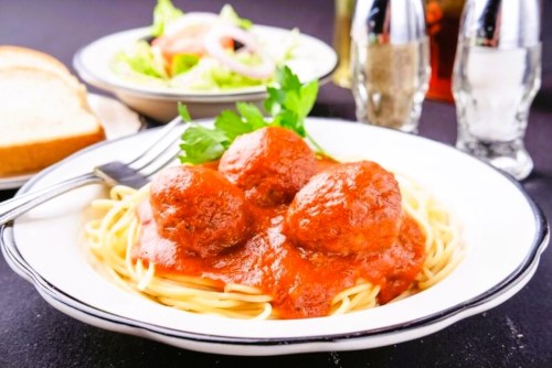 A plate of spaghetti topped with meatballs and marinara sauce, with a side salad and bread in the background.