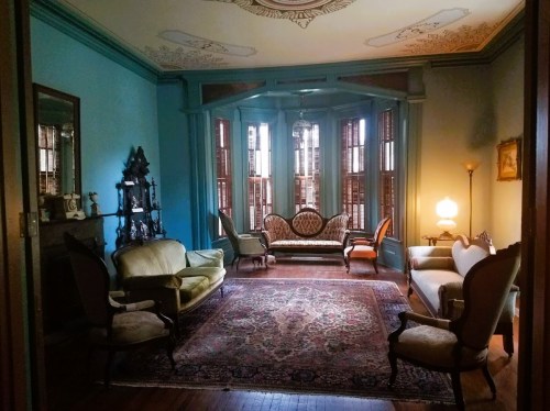 A vintage living room with ornate furniture, a patterned rug, and large windows, featuring soft lighting and decorative details.