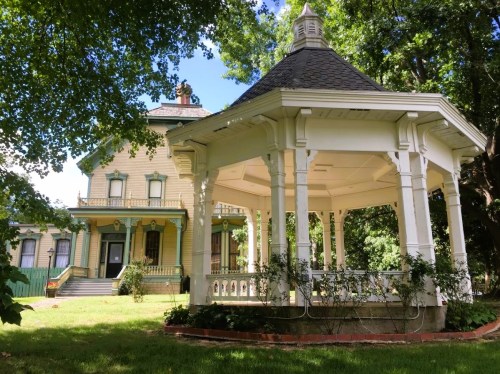 A historic yellow house with green accents, next to a white gazebo surrounded by lush greenery.