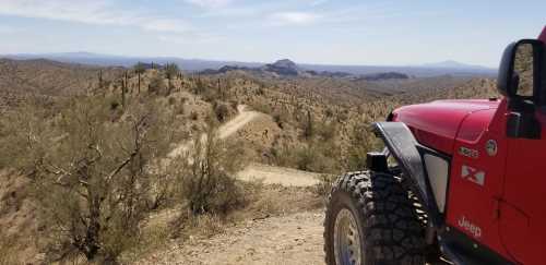 A red Jeep parked on a desert trail, overlooking a mountainous landscape with cacti and blue skies.